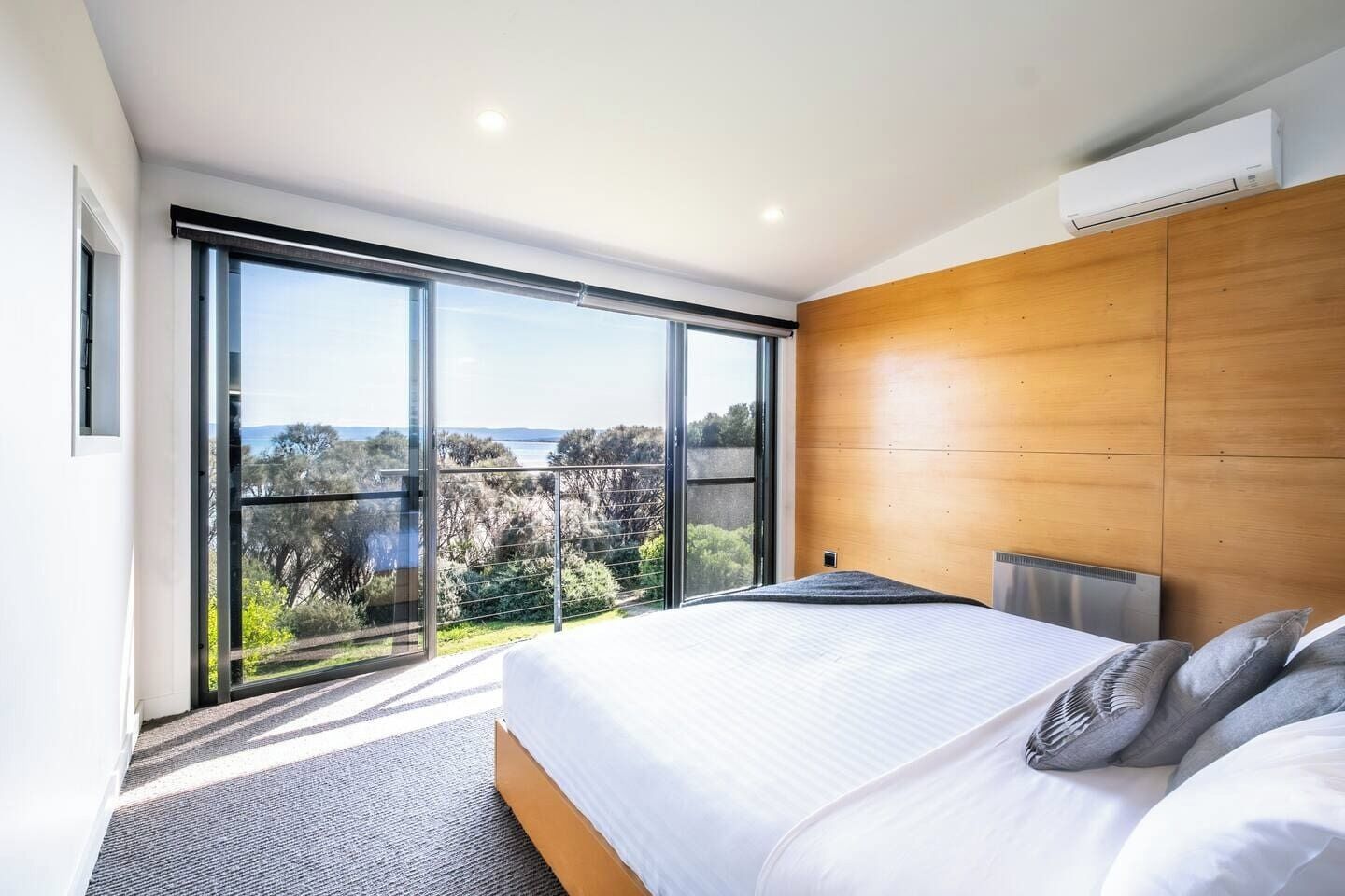 A modern bedroom with a large bed, a wooden headboard, and a view of the outdoors through floor-to-ceiling windows.
