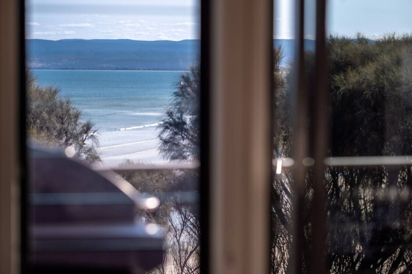 Vista de una playa a través de una ventana, con árboles en primer plano y un cielo despejado.
