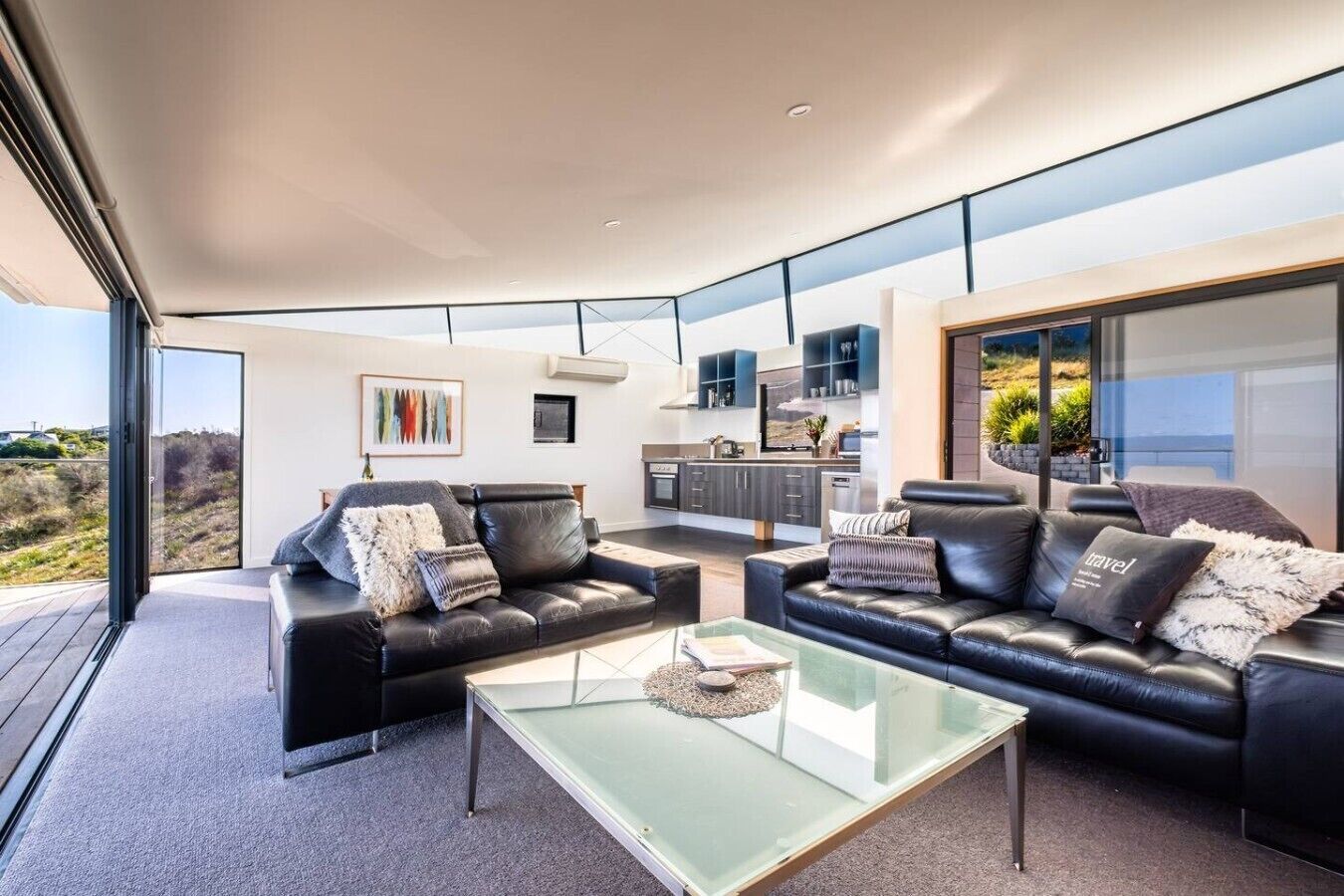 A modern living room with two black leather sofas, a glass coffee table, and a view of the kitchen and outdoor landscape through large windows.