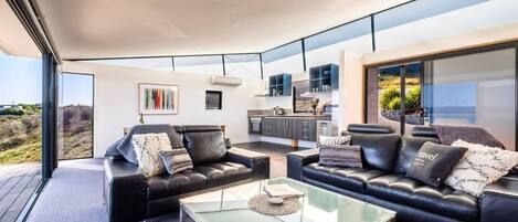A modern living room with two black leather sofas, a glass coffee table, and a view of the kitchen and outdoor landscape through large windows.