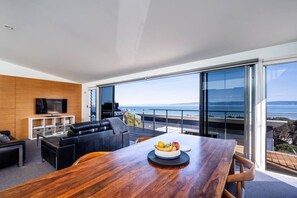 A modern living room with a wooden dining table, a flat-screen TV, and a balcony with a view of the sea. - Footprints 1 ~ Ocean Views - Beachfront - Sunsets (Coles Bay)