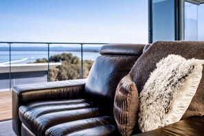 A modern lounge area with a leather sofa and a fluffy pillow, overlooking a scenic view through a glass railing. - Footprints 1 ~ Ocean Views - Beachfront - Sunsets (Coles Bay)
