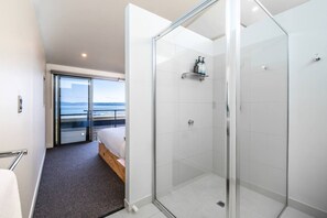 A modern hotel room with a glass shower enclosure, a bed with a view of the sea, and a balcony. - Footprints 1 ~ Ocean Views - Beachfront - Sunsets (Coles Bay)