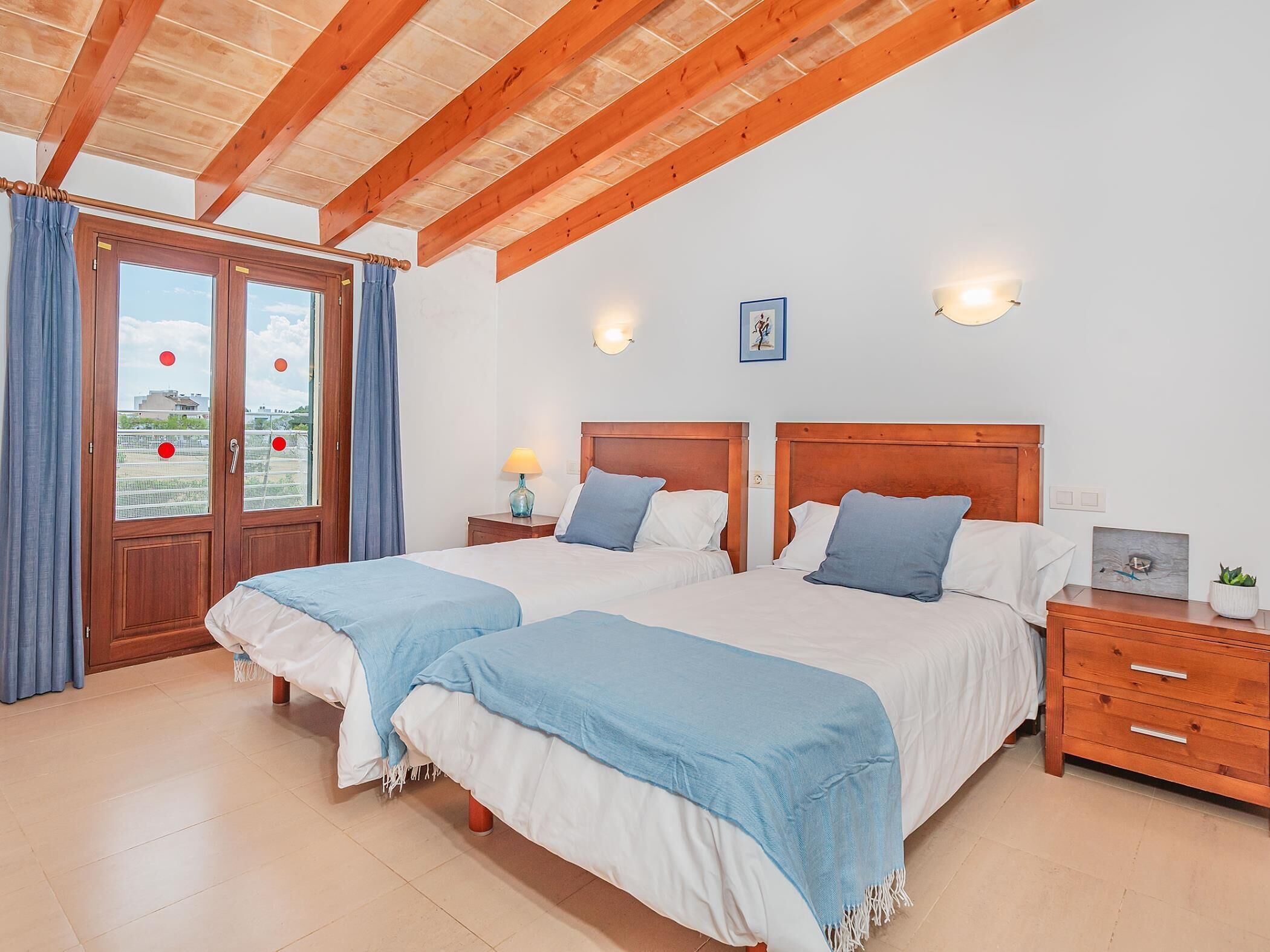 A bedroom with two beds, wooden ceiling beams, a window with curtains, a bedside table with a lamp, and a small plant on a nightstand.