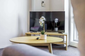 Une pièce avec un téléviseur posé sur un meuble, une table ronde ornée d’une plante en pot et une télécommande sur la table.