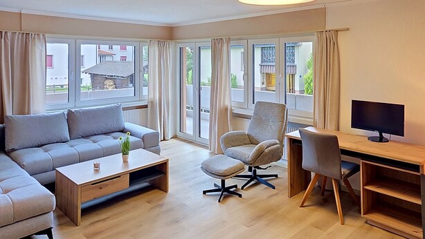 A modern living room with a sofa, armchair, coffee table, and a desk with a computer. Large windows offer a view of the outside.