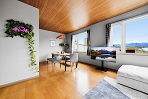 Una habitación moderna con techo de madera, zona de comedor con mesa y sillas, cama y televisor que muestra un paisaje.