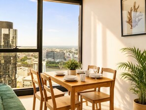 Een eetruimte met een tafel voor vier personen, een kamerplant en stadszicht door grote ramen.