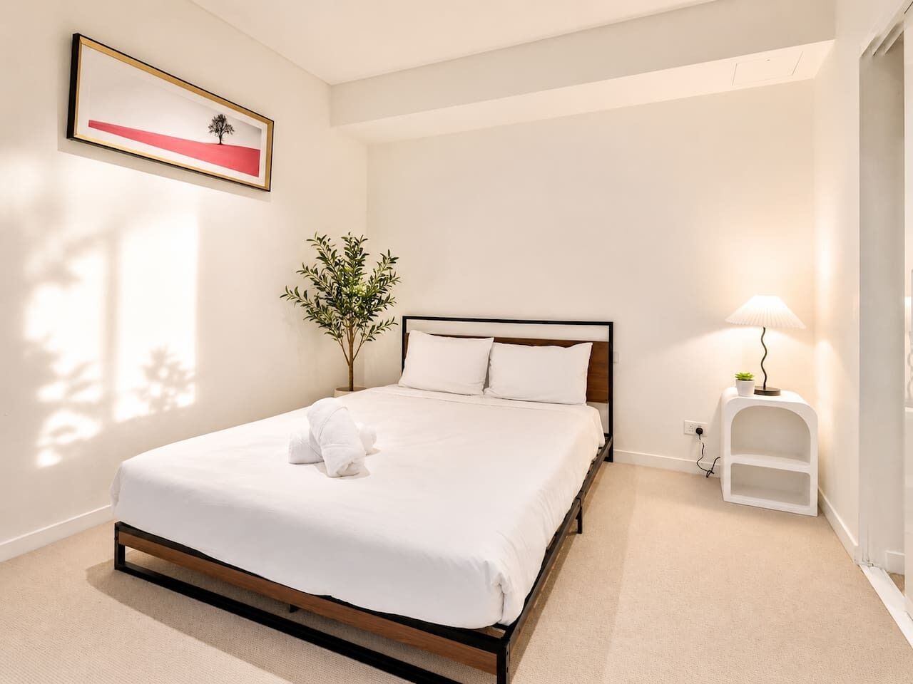 Een net opgemaakt bed met witte lakens, een bedlampje, een klein wit nachtkastje en een lijst met een foto aan de muur.