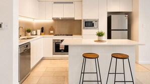 A modern kitchen with white cabinets, stainless steel appliances, a central island with stools, and a dishwasher. - CBD Luxury 2B w/ Parking & Sky View (Brisbane City)
