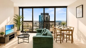 A modern living room with a large window, a green sofa, a dining table, and a TV. - CBD Luxury 2B w/ Parking & Sky View (Brisbane City)