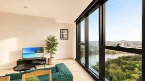 A modern living room with a large window, a flat-screen TV, a potted plant, and a green sofa. - CBD Luxury 2B w/ Parking & Sky View (Brisbane City)