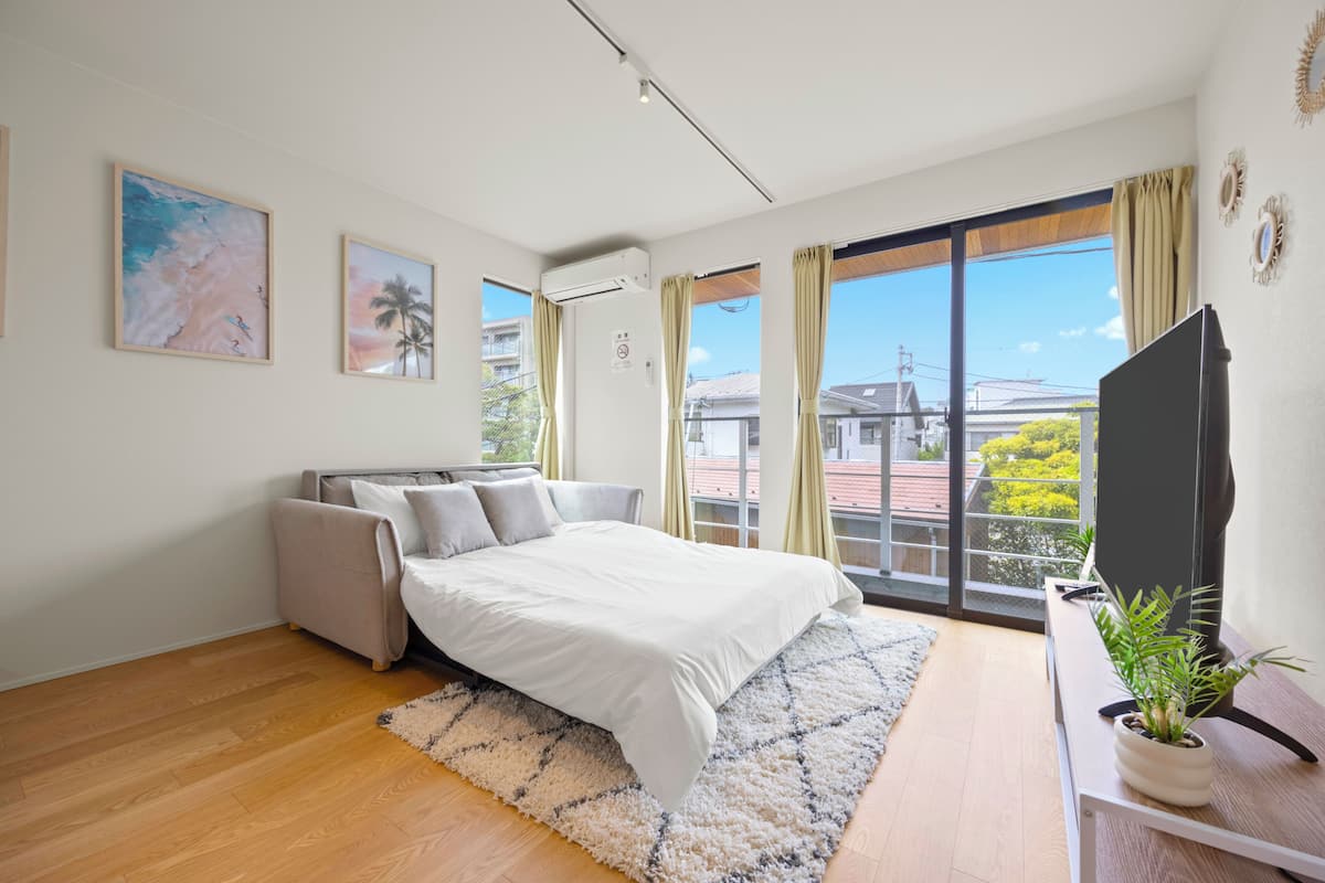A bedroom with a bed, a sofa, a TV, a plant, and large windows.