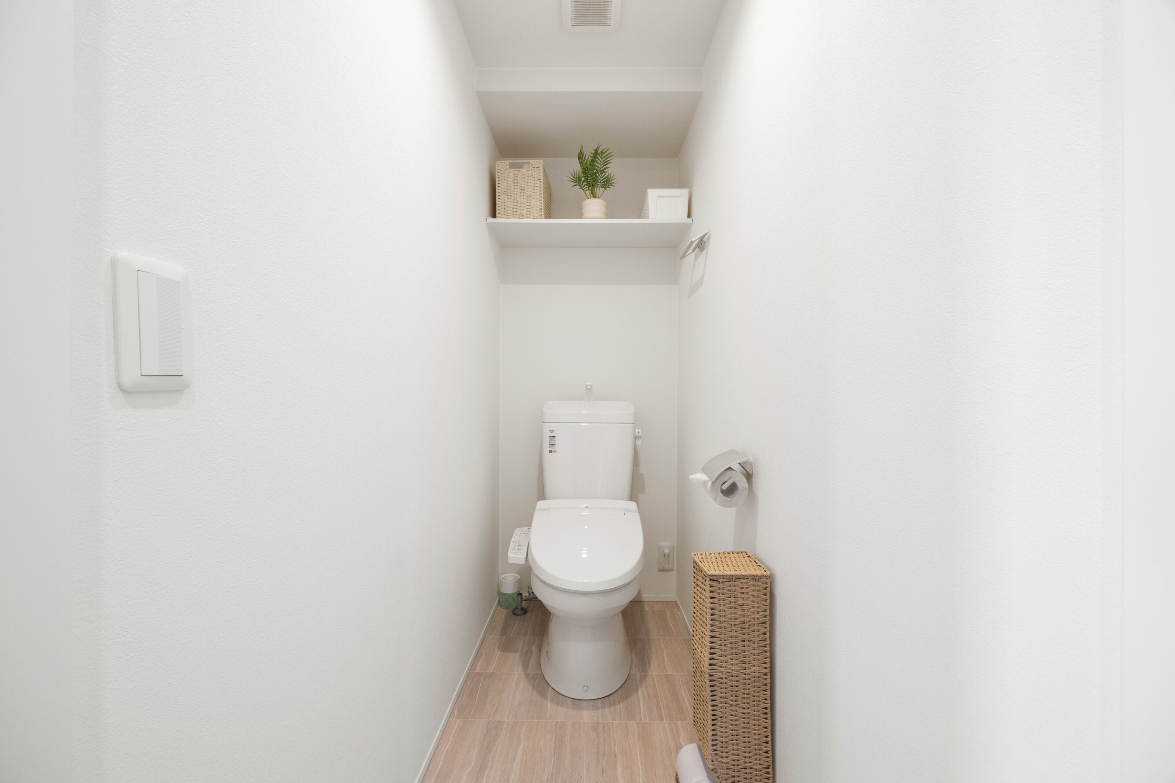 Un baño estrecho con inodoro, un estante con una planta y una cesta, e interruptor de luz en la pared.