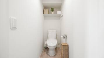 Un baño estrecho con inodoro, un estante con una planta y una cesta, e interruptor de luz en la pared.