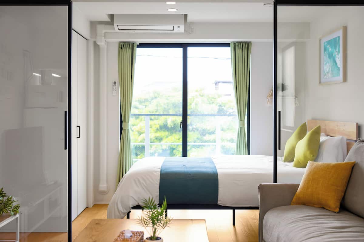 A modern hotel room with a bed, a sofa, a small table with a plant, and a large window with curtains.