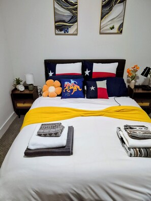 En snyggt bäddad säng med ett gult överkast, en kudde med en Texas-flagga och en kudde med ett motiv från vilda västern. Det finns två sängbord med en lampa, en klocka och en liten växt.