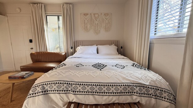 Een slaapkamer met een bed, een bruine stoel, een klein tafeltje en een wanddecoratie van macramé.