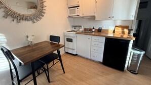 Een compacte keuken met witte kastjes, een houten eettafel, een zwarte stoel, een magnetron, een fornuis en een prullenbak.