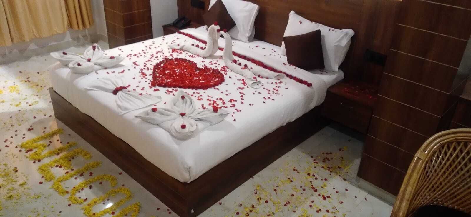 A neatly made bed with rose petals and swan-shaped towels, a heart-shaped arrangement of red petals, and a 'WELCOME' sign on the floor.