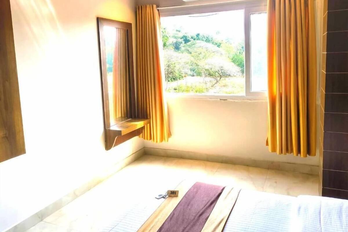 A bedroom with a bed, an air conditioning unit, a window with curtains, and a view of greenery outside.