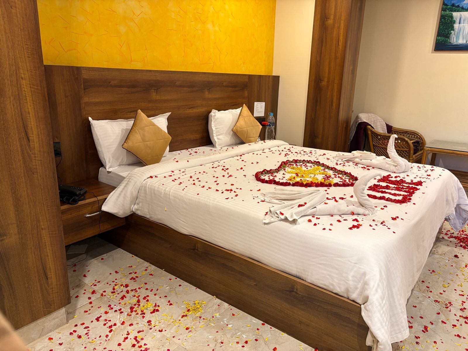 Una habitación de hotel con cabecera de madera, cama con ropa de cama blanca y una decoración con forma de corazón hecha de pétalos de rosa.