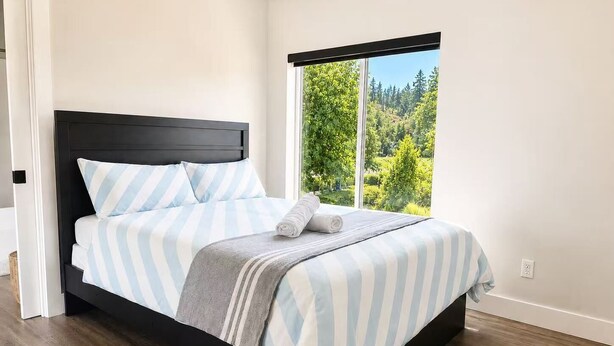 A bedroom with a large bed, a view of greenery through a window, and a wooden floor. - Modern 2 Bedroom Lakeview Suite (Kelowna)