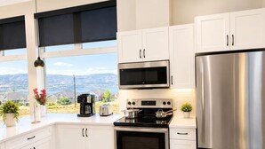 A modern kitchen with white cabinets, stainless steel appliances, a large window with a view, and a potted plant on the counter. - Modern 2 Bedroom Lakeview Suite (Kelowna)