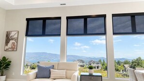 A living room with a sofa, a side table, and a view of a residential area through large windows. - Modern 2 Bedroom Lakeview Suite (Kelowna)