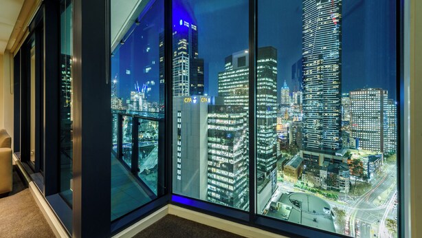 A modern hotel room with large floor-to-ceiling windows offering a nighttime view of a cityscape with illuminated skyscrapers. - Exclusive Stays - SouthbankONE (Southbank)