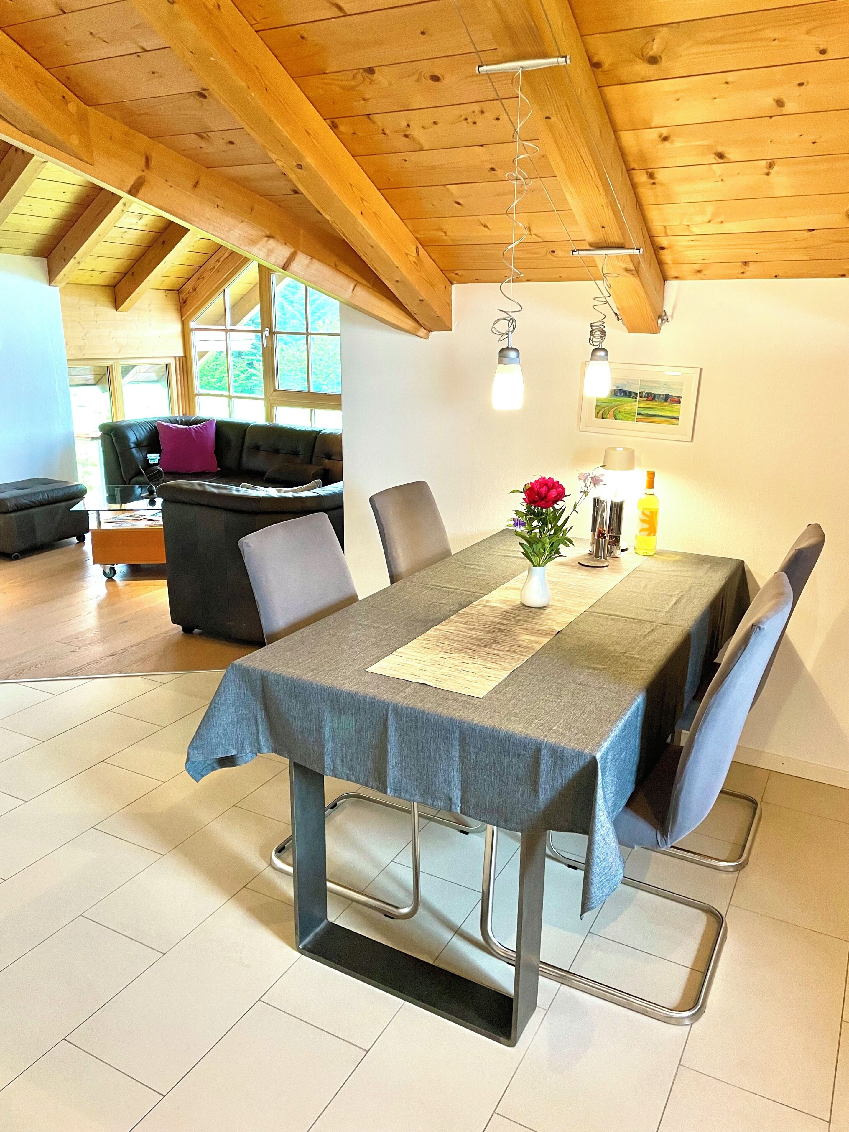 A dining area with a table set for four, wooden beams on the ceiling, a sofa in the background, and a window with a view of greenery.