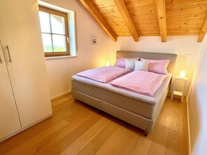 Ein Schlafzimmer mit einem Bett, Holzbalken an der Decke, einem Fenster und einem Nachttisch mit Lampe.