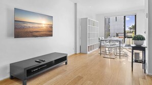 Un salon moderne doté d’un grand téléviseur, d’un meuble de rangement noir pour appareils multimédias, d’un coin repas avec des chaises transparentes et d’une bibliothèque.