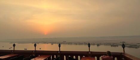 An outdoor seating area with tables and chairs, overlooking a body of water with boats and a sunset in the distance.