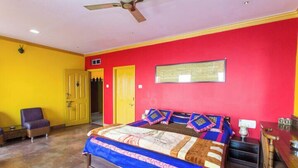 A bedroom with a bed, a chair, a desk, a ceiling fan, and a door. - Ganpati Guest House (Varanasi)