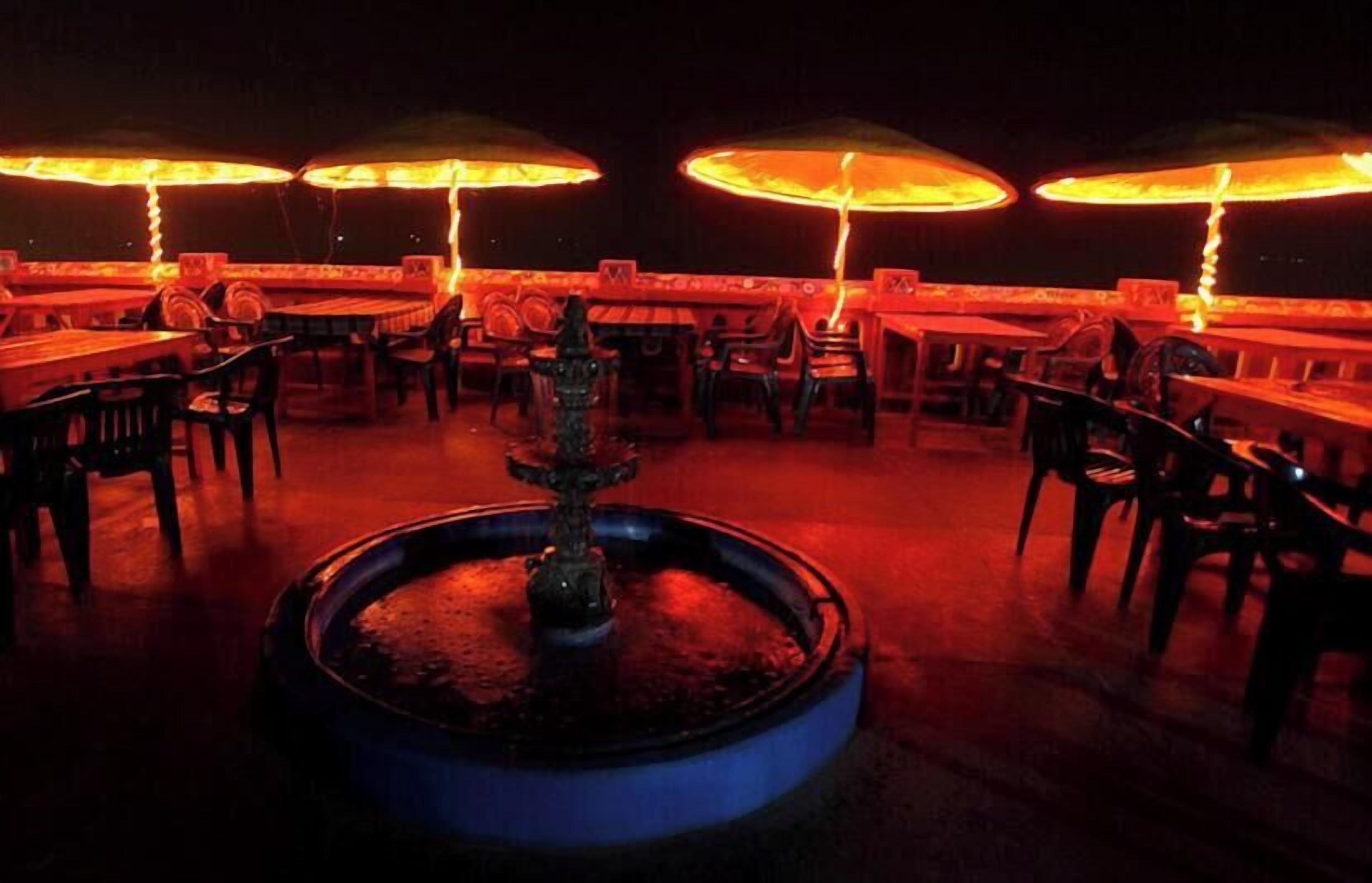 Un espace de restauration en plein air avec des tables et des chaises, une fontaine centrale et un éclairage assuré par des parasols.