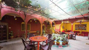 A covered outdoor seating area with tables and chairs, potted plants, and a mural on the wall. - Ganpati Guest House (Varanasi)