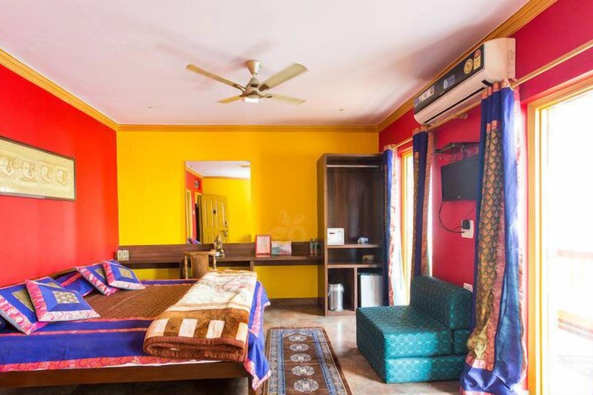 Una habitación con dos camas, ventilador de techo, aire acondicionado, estantería y ventana con cortinas.