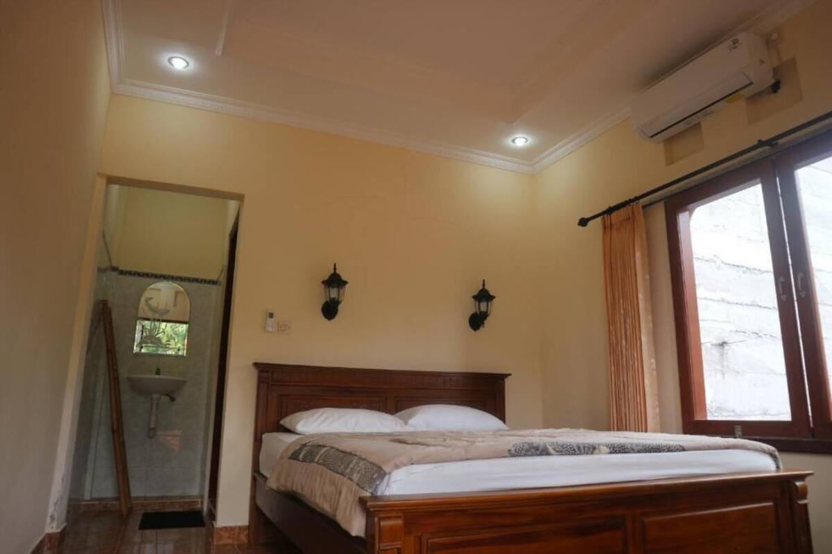 A bedroom with a wooden bed, a window with curtains, a wall-mounted air conditioner, and a mirror in the corner.