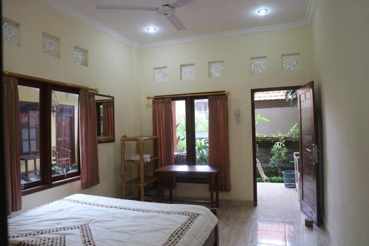 A bedroom with a bed, a desk, a chair, a ceiling fan, and a view of a garden.