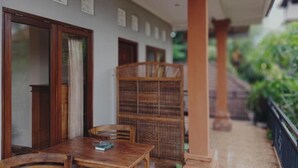 A covered outdoor seating area with a table and chairs, a bookshelf, and a view of greenery. - Sudana Homestay Ubud (Ubud)