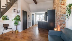 A spacious living area with a wooden floor, a staircase, a kitchen with a brick wall, and a blue sofa. - Two-bedroom Apartment by the River (Antwerp)