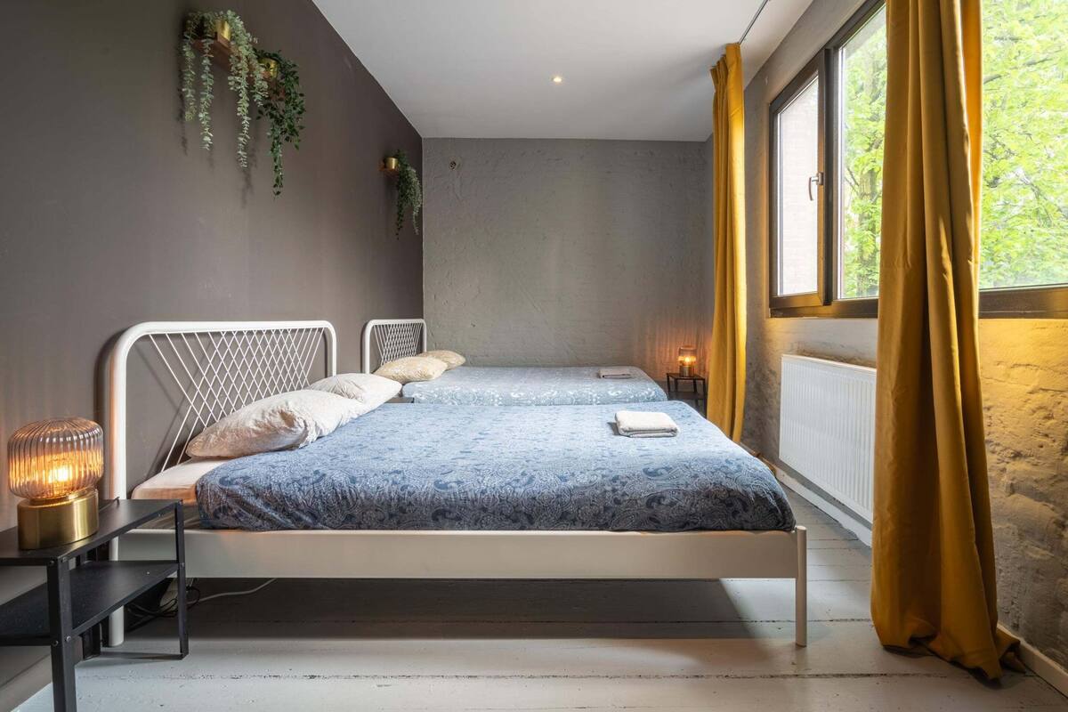 Una cama doble con una colcha estampada, una mesita de noche con lámpara y una ventana con cortinas.