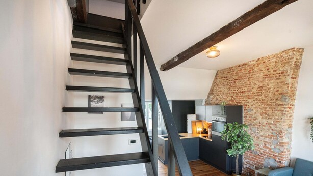A staircase with black railings leading to a kitchen area with a brick wall and a blue sofa. - Two-bedroom Apartment by the River (Antwerp)