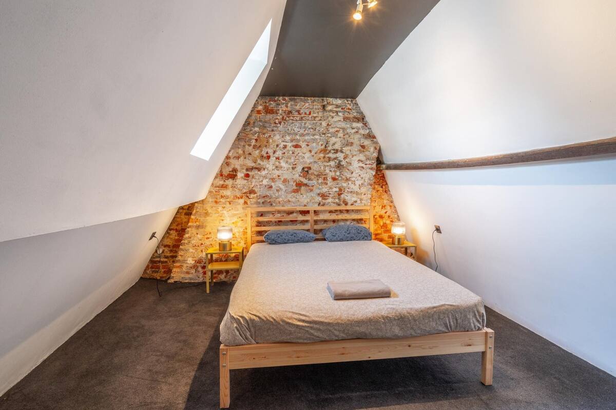 Un dormitorio con techo inclinado, una cama, dos mesitas de noche con lámparas y una pared de ladrillo.