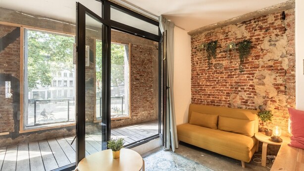 A room with a brick wall, a sofa, a table, and a window with a view of the street. - The Industrial Apartments 2bbknsinternet (Antwerp)