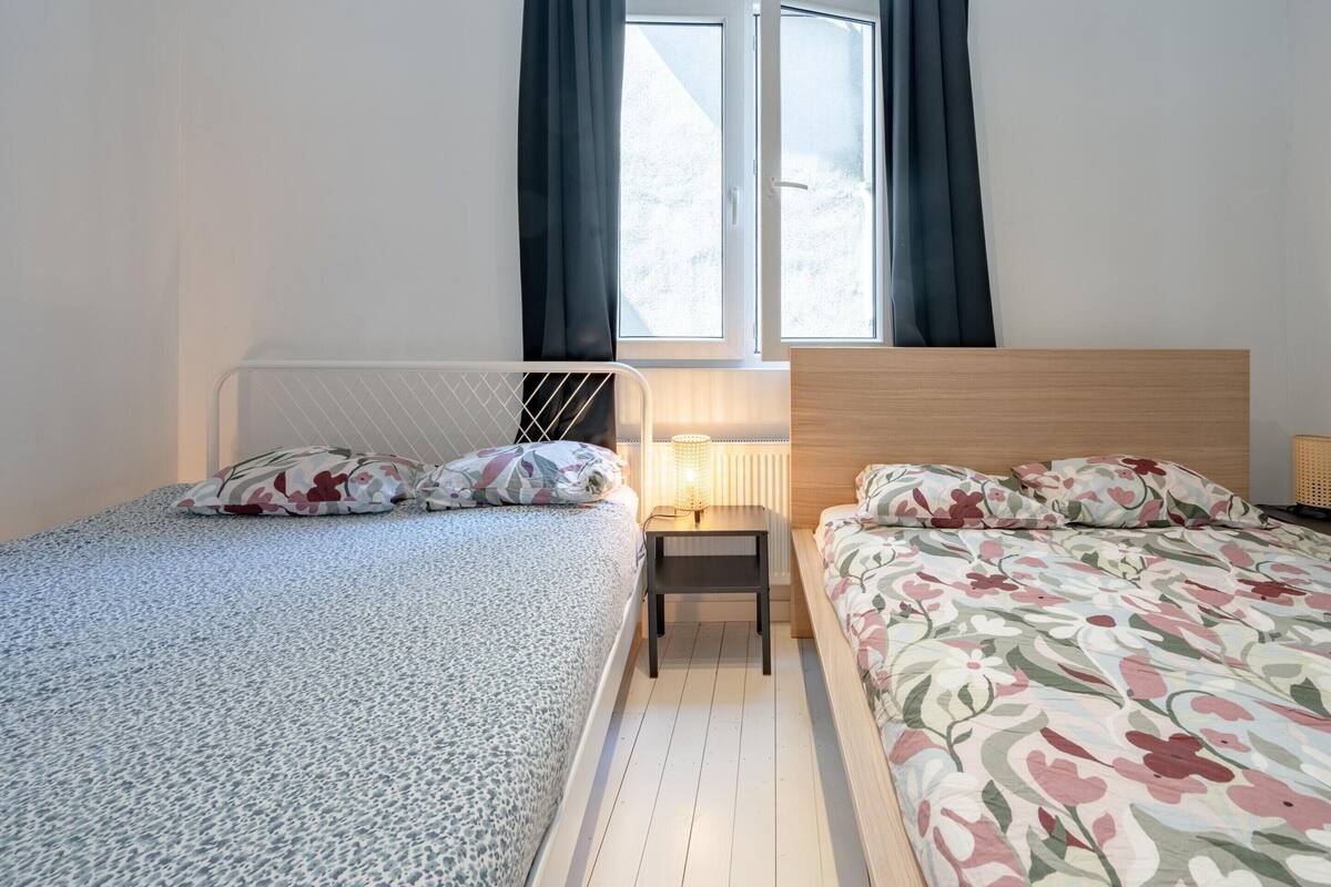 Un dormitorio con dos camas, una mesita de noche, un ventanal con cortinas y un radiador.