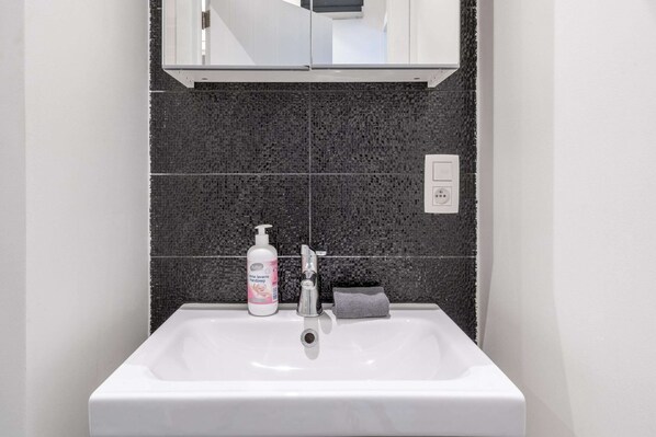 Een badkamer met een wit wastafelblad, een donkere betegelde achterwand, een spiegel boven de wastafel en een handzeepfles.
