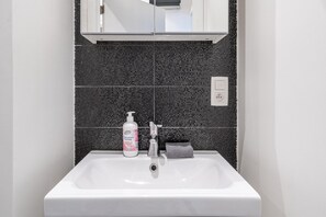 Un baño con un lavabo blanco, un salpicadero de azulejos oscuros, un espejo sobre el lavabo y una botella de jabón para manos.