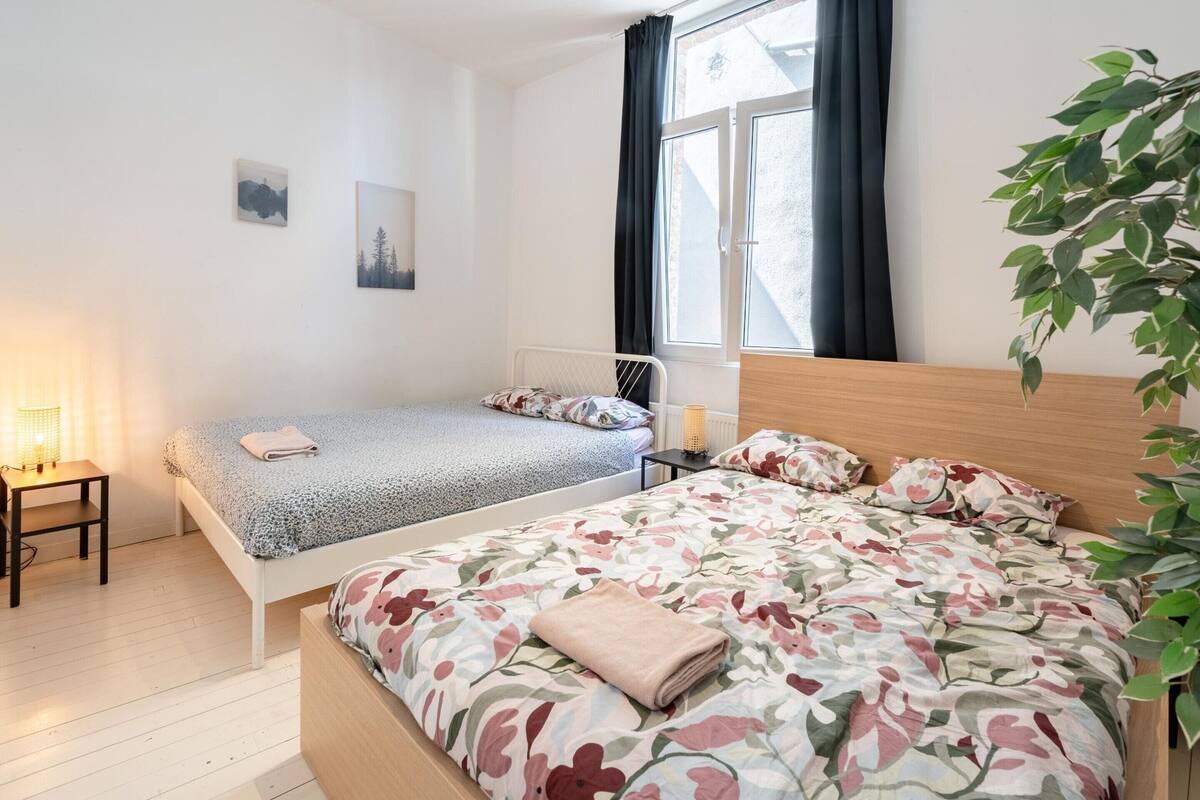 Un dormitorio con dos camas, una mesita de noche con lámpara, un radiador y una ventana con cortinas.